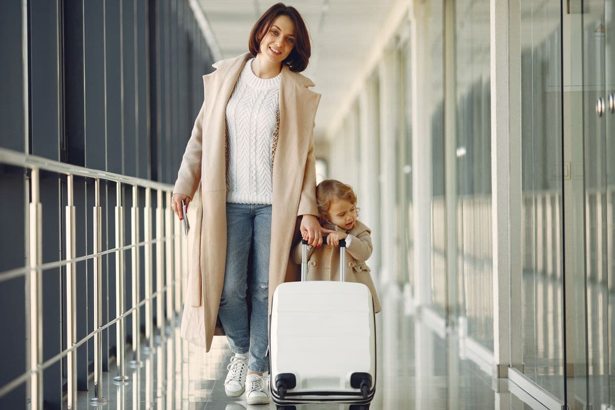Mother and young daughter walking through an airport corridor with a rolling suitcase